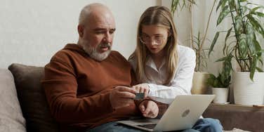 young woman showing her older father how to use a bank card and computer at home