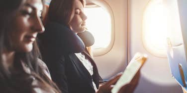 woman sleeping on flight sitting beside window while woman sitting beside her reads e-book
