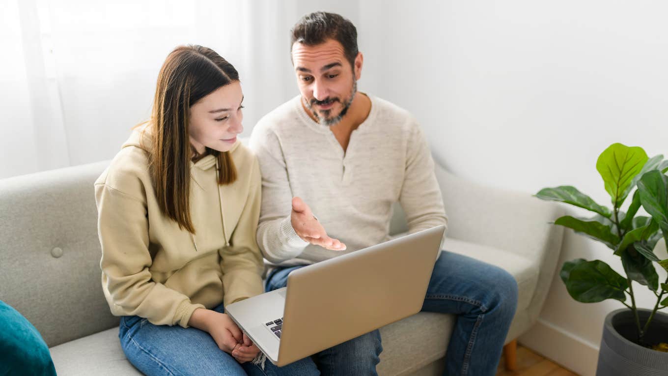 father and teen daughter sitting on couch and talking while looking at laptop