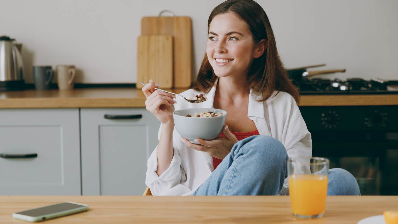 young woman smiling while eating a bowl of cereal at her dining table