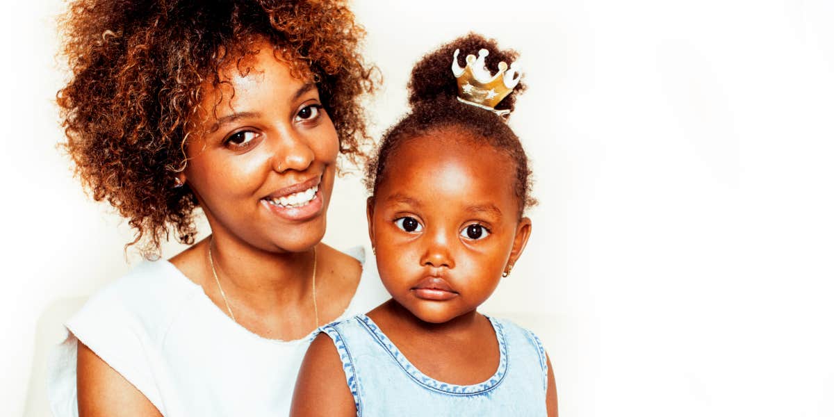 Black mother with natural hair smiles, her daughter with hair pulled into a bun looks serious