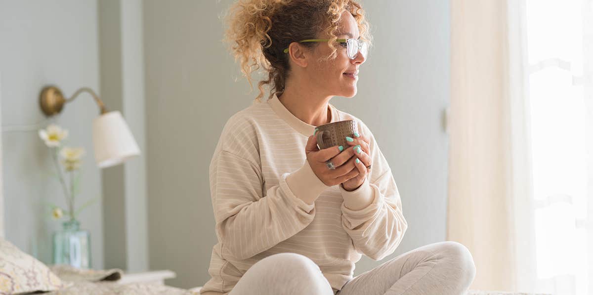 Beautiful caucasian young woman with curly hair and eyeglasses sitting on bed and looking away while drinking coffee in morning