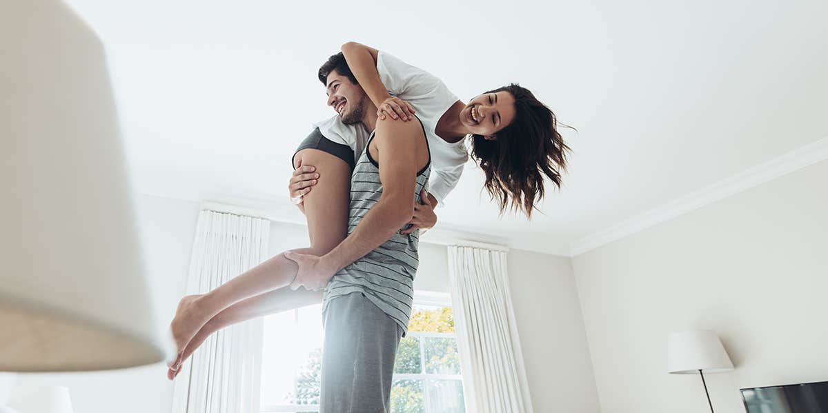 There's Only 4 Types Of Love Attachment Styles — Which Is Yours? Young man standing on bed and carrying his girlfriend on his shoulder. Couple in playful mood in bedroom.