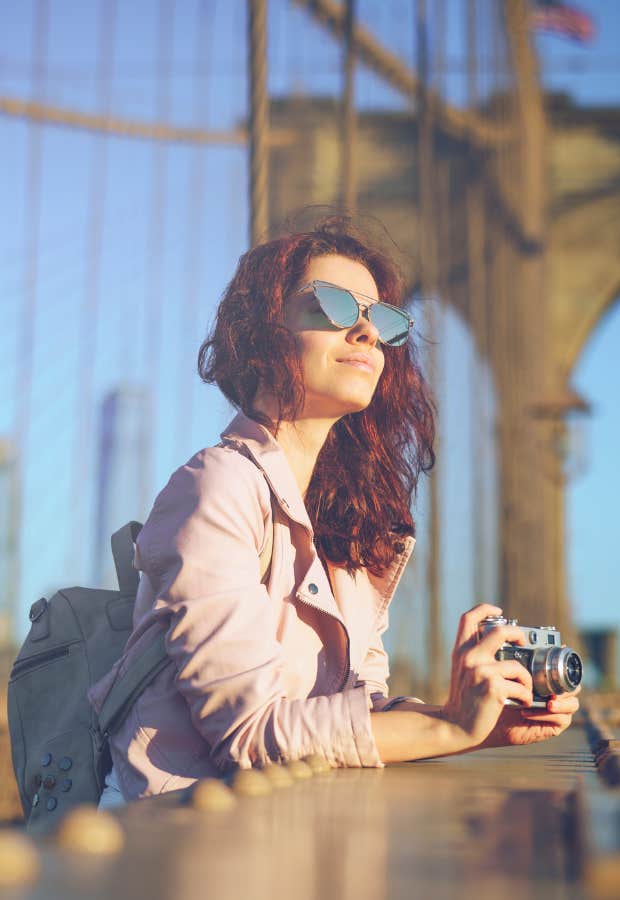 A woman holds a camera and stands in the middle of the Brooklyn Bridge.