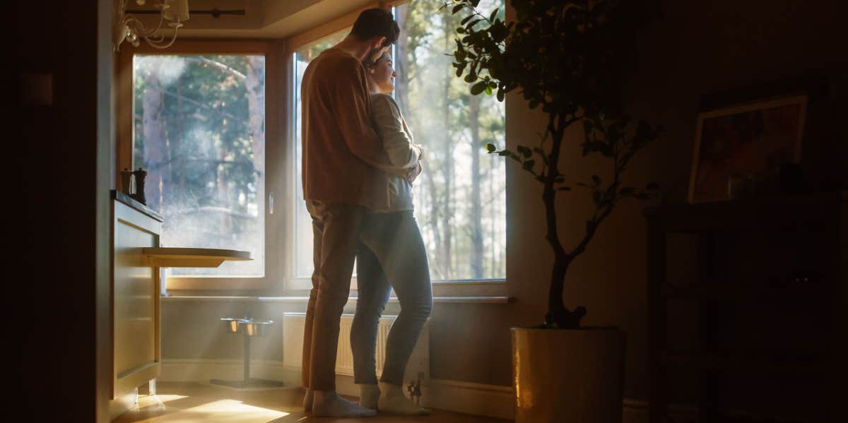 Woman Watching Out of Window Male Partner Embraces Her From Behind