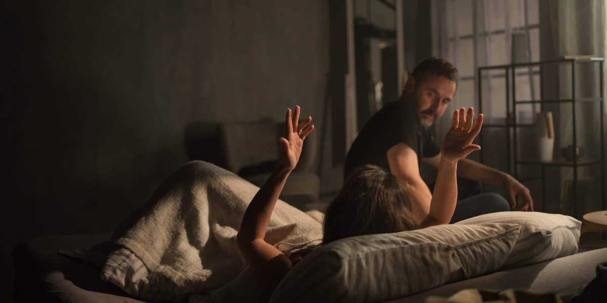 boyfriend sitting on bed introgating girlfriend, who throws hands in air