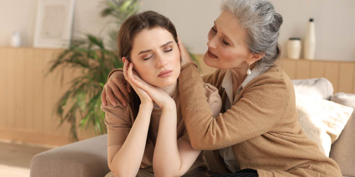 Middle aged mother in law is embracing her daughter in law, comforting upset frustrated daughter in law with closed eyes, touching cheeks, sitting on couch at home. 