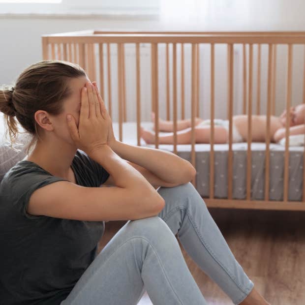 Woman sits on the floor next to a baby crib with her hands on her face, seemingly in distress