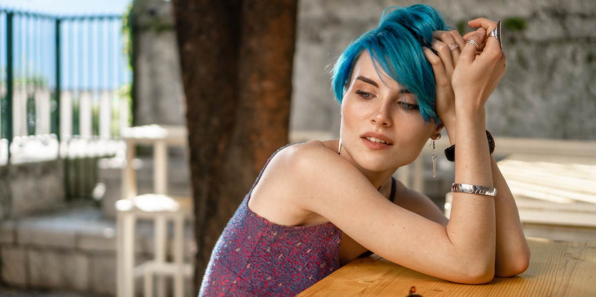 A girl in a purple dress and blue hair sits at a wooden table on a cafe terrace with paved walls