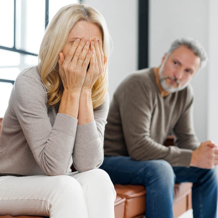 frustrated blonde woman covered face with palms and crying while sitting bside sad grey-haired mature man on couch