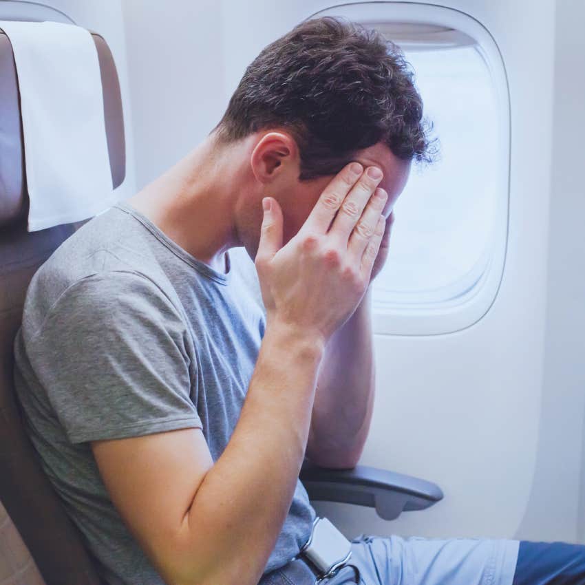 male passenger with hands on face suffering from flight anxiety while sitting next to window on plane