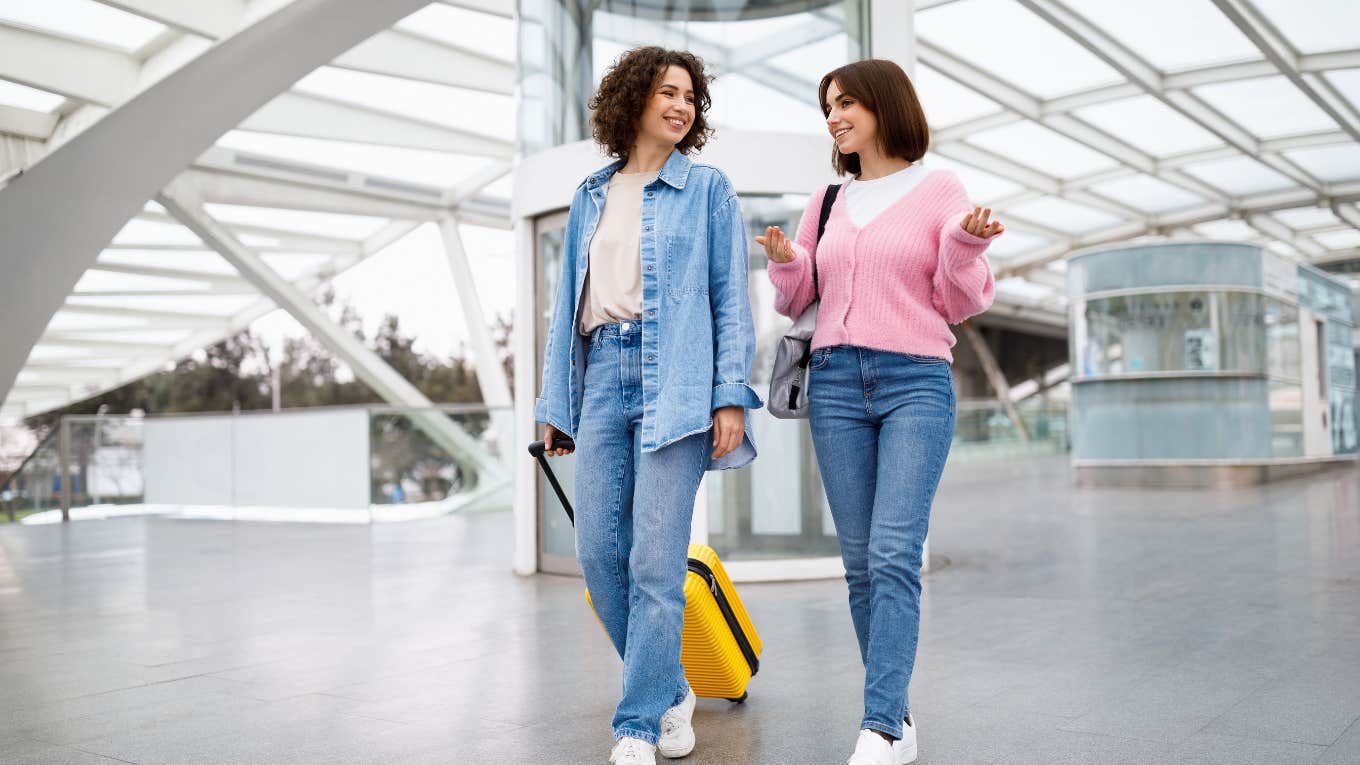 two smiling women walking through airport together with luggage