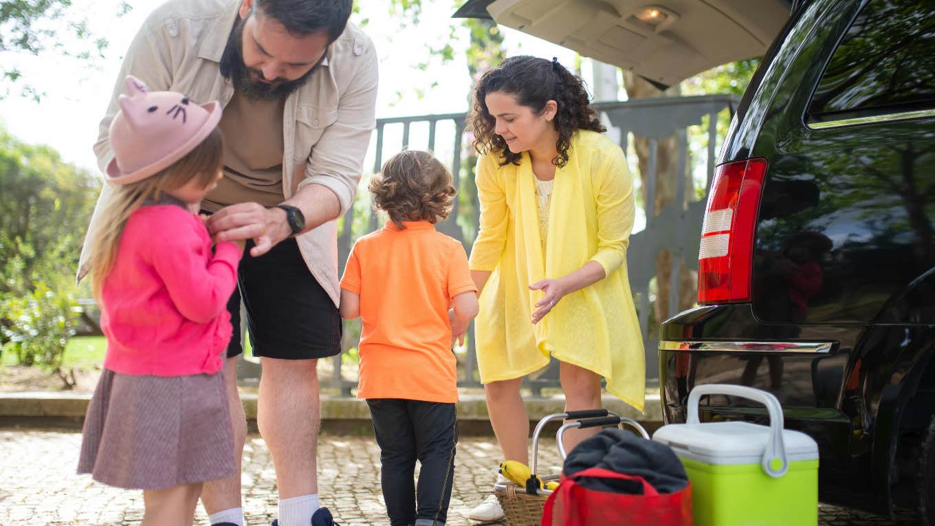 Dad Stands By While His Wife Trips Over Unpacking Bags And Their Two Kids From The Car family unpacking their car