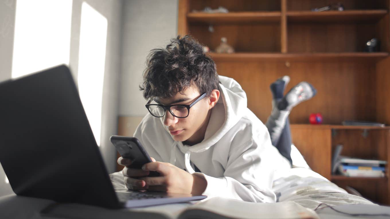 Teen Forced To Pay For His Own Car, Gas & Bills Now Told He Needs To Pay 15% Of His Income To His Parents For Rent young boy studying while lying on the bed using computer and smartphone