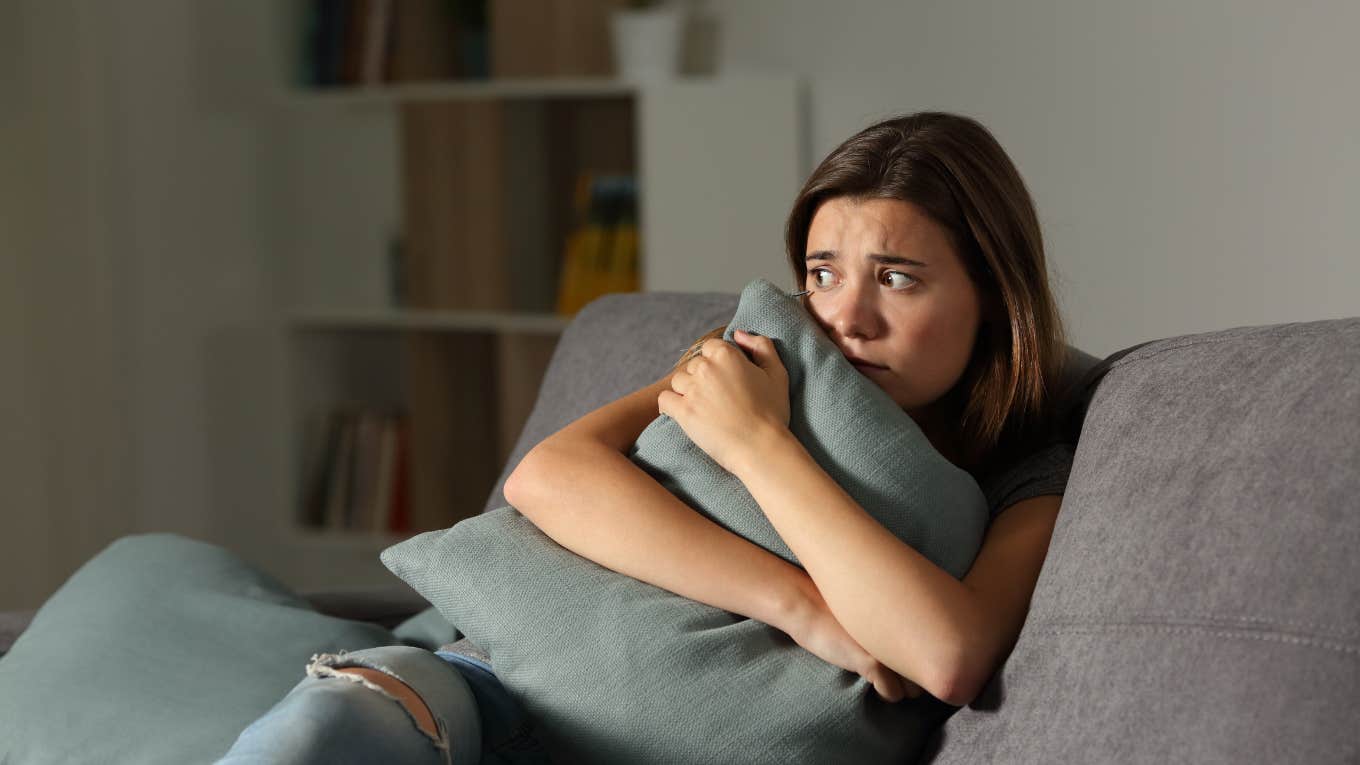 Woman Found Squatter Living In Her Bedroom For 4 Months Without Her Realizing Scared woman at home embracing pillow sitting on a couch in the living room at home
