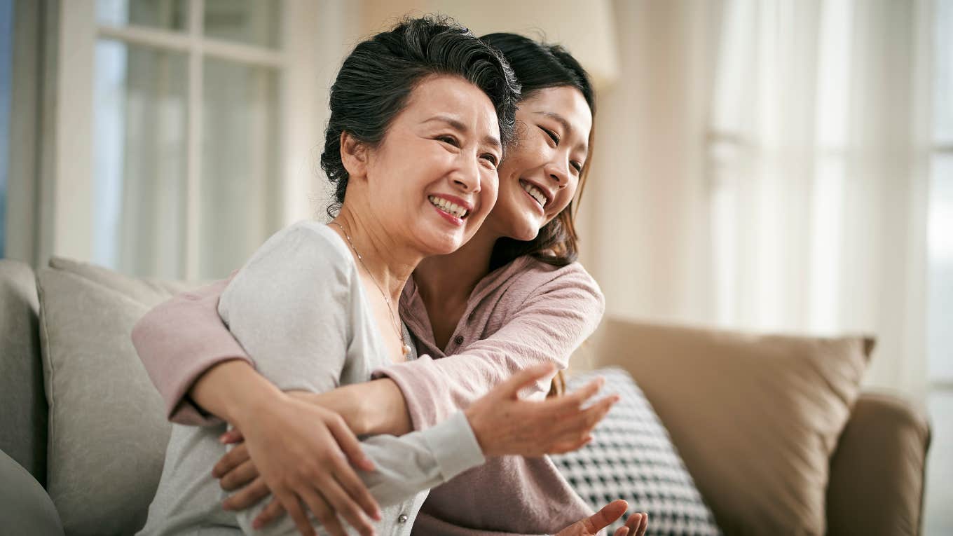 loving asian adult daughter sitting on couch at home hugging senior mother happy and smiling
