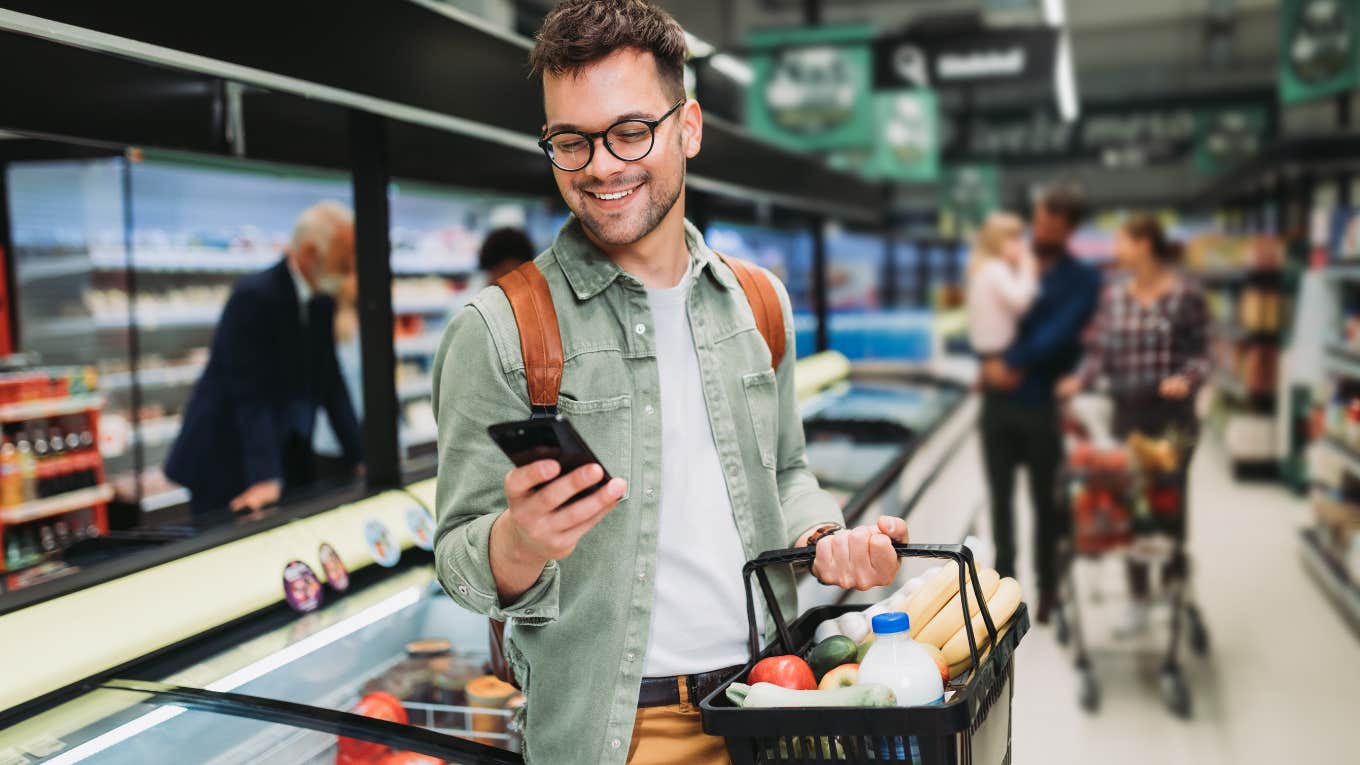 Groceries Are Sadly The New ‘Splurge’ Item For Millennials and Gen Z Gen-Z man grocery shopping while on his phone.