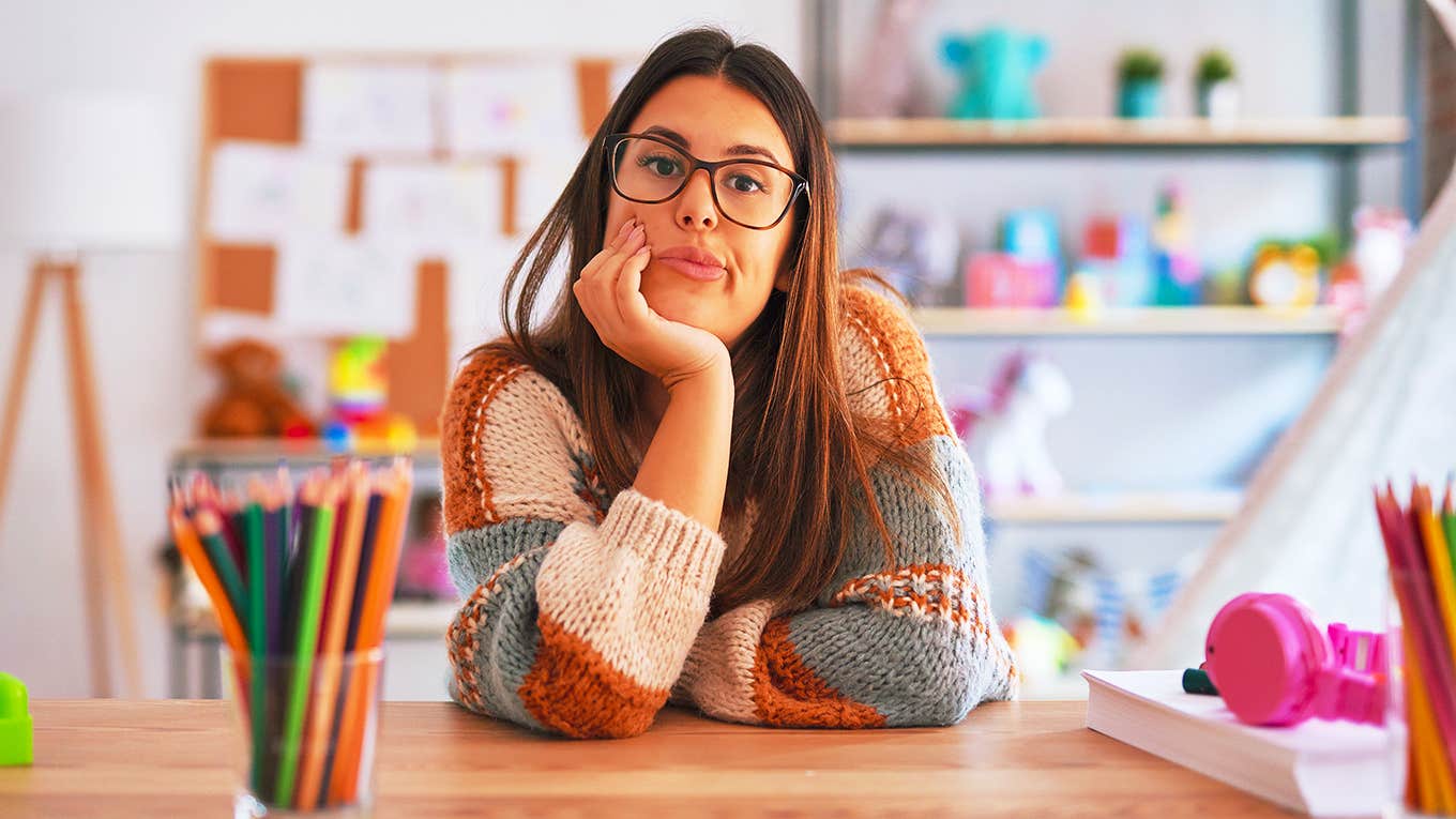 High School Teacher Stresses Her Impatience With The Learned Helplessness Of Her Students teacher woman wearing sweater and glasses sits at desk looking frustrated and annoyed with crossed arms.