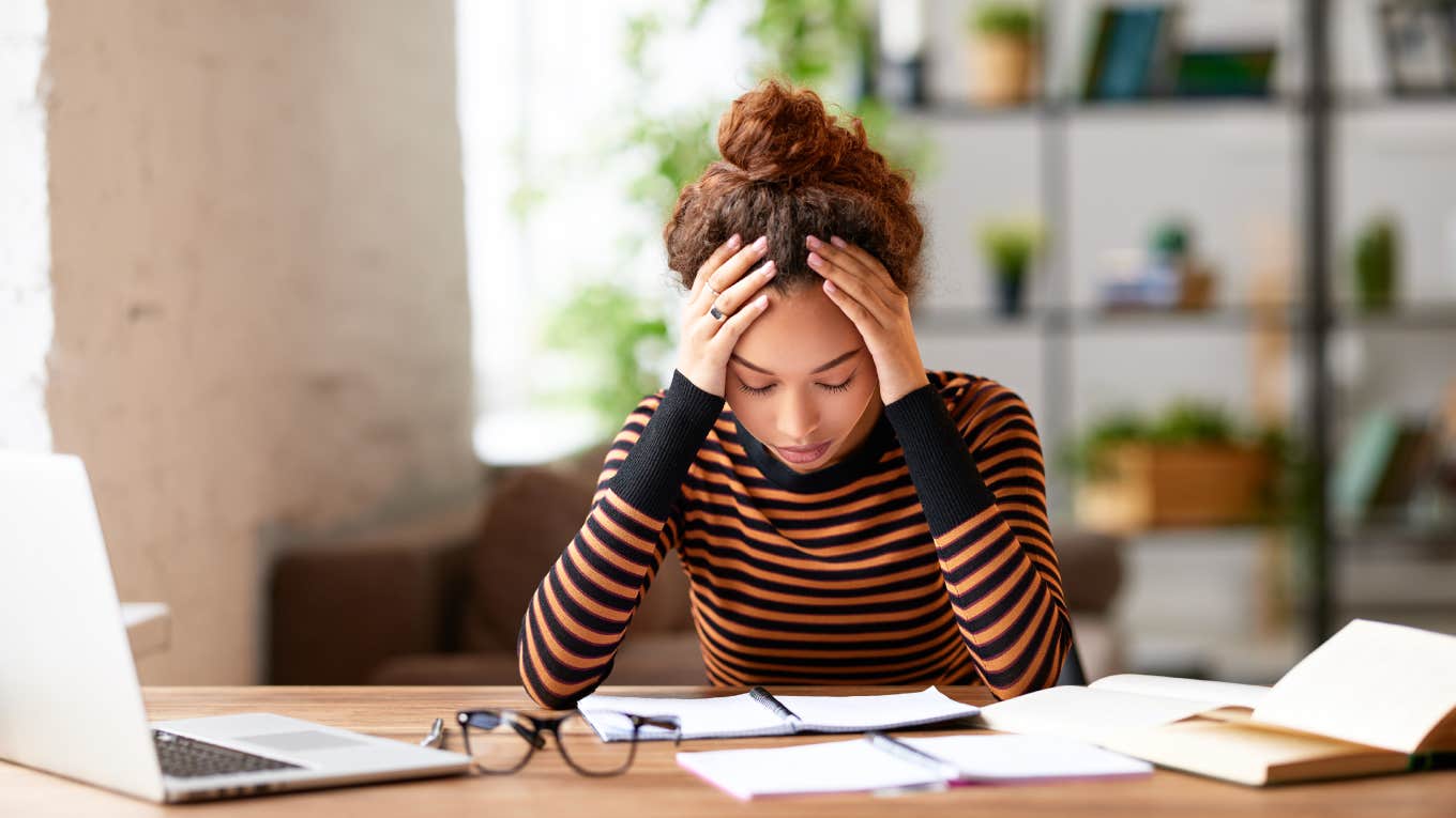 25-Year-Old Who Works 7 Days A Week & Goes To Grad School Can't Afford To Quit Her Jobs And Still Make Rent woman holding head in hands and feeling demotivated while sitting at her home office and working remotely on laptop.