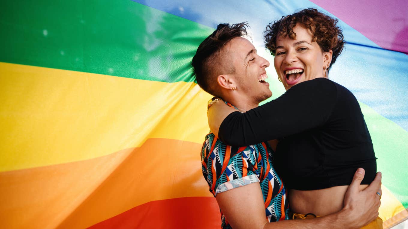 The Unique Phrase That Explains Why Time ‘Feels Different’ To Queer And Trans People queer couple embrace each other in front of pride flag