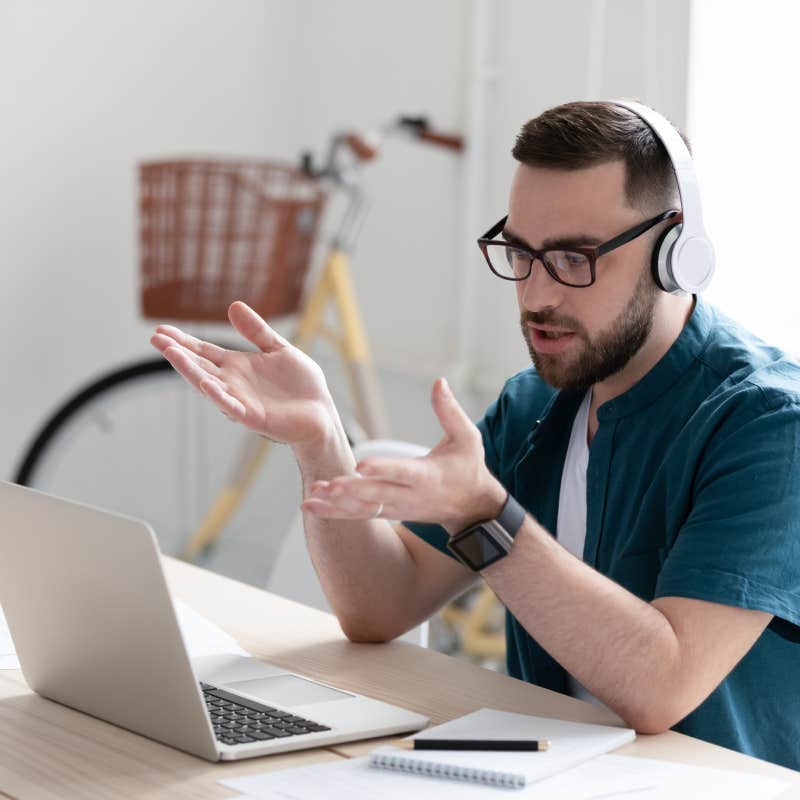 focused young businessman in eyewear wearing headphones, holding video call with clients on laptop