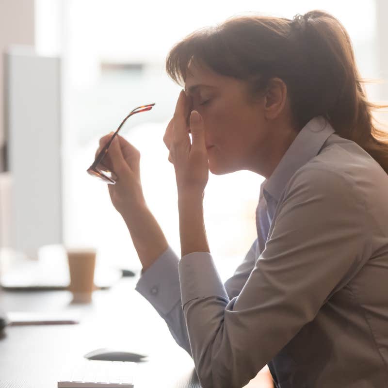 tired female worker sitting at desk with glasses off rubbing at eye