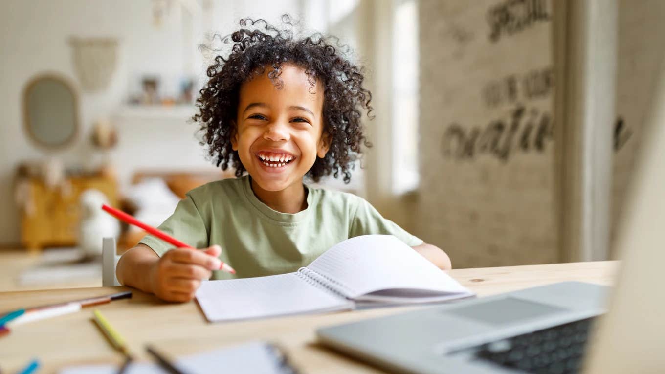 Mom Told Her Son Needs To Have An Evaluation Because of His Low IQ But His Teachers Disagree smiling little boy sitting in classroom with book open and pencil in hand
