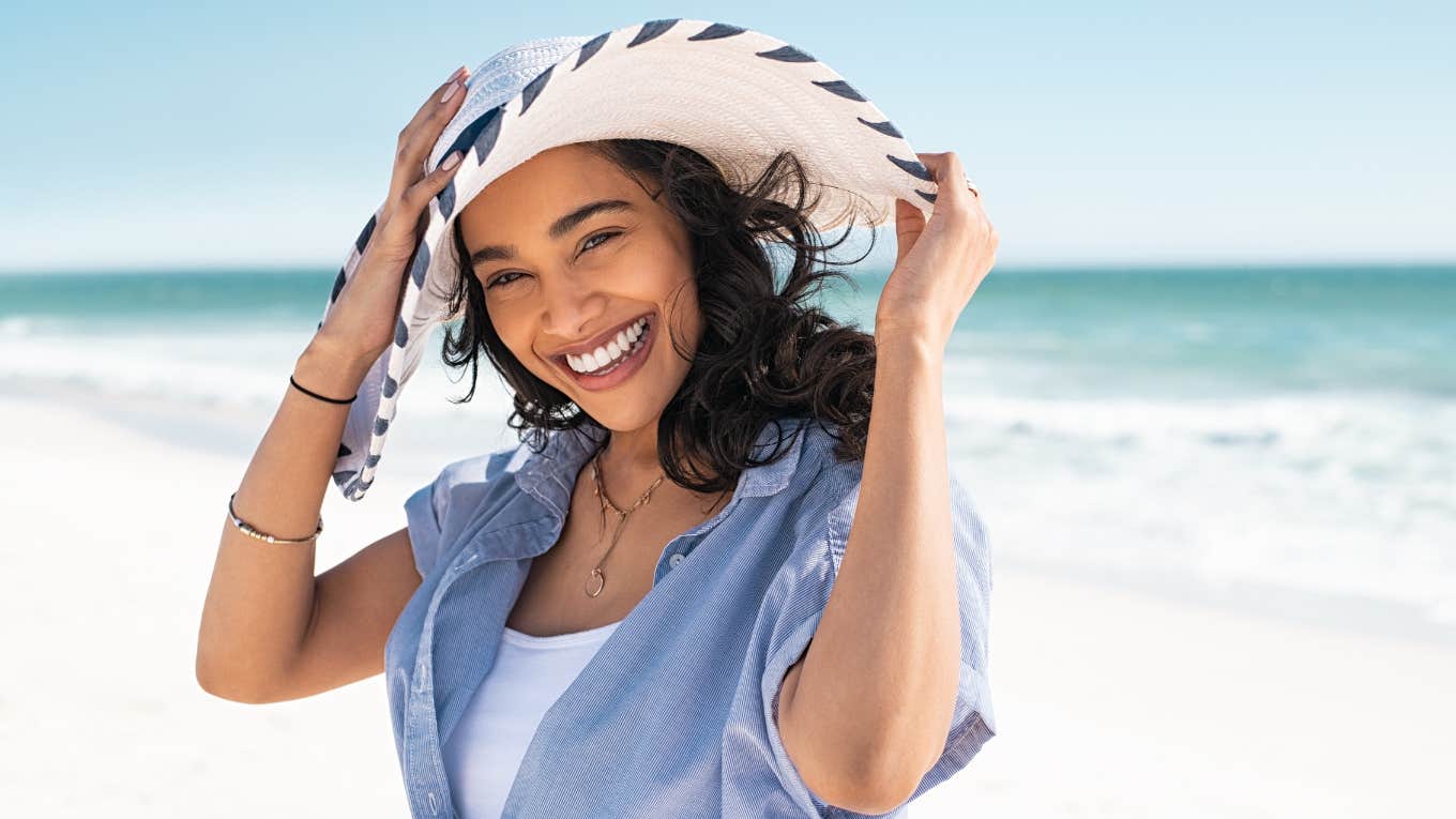 A 2024 Poll Reveals Exactly How Much Money You Need To Be Happy Smiling woman wearing sun hat on the beach