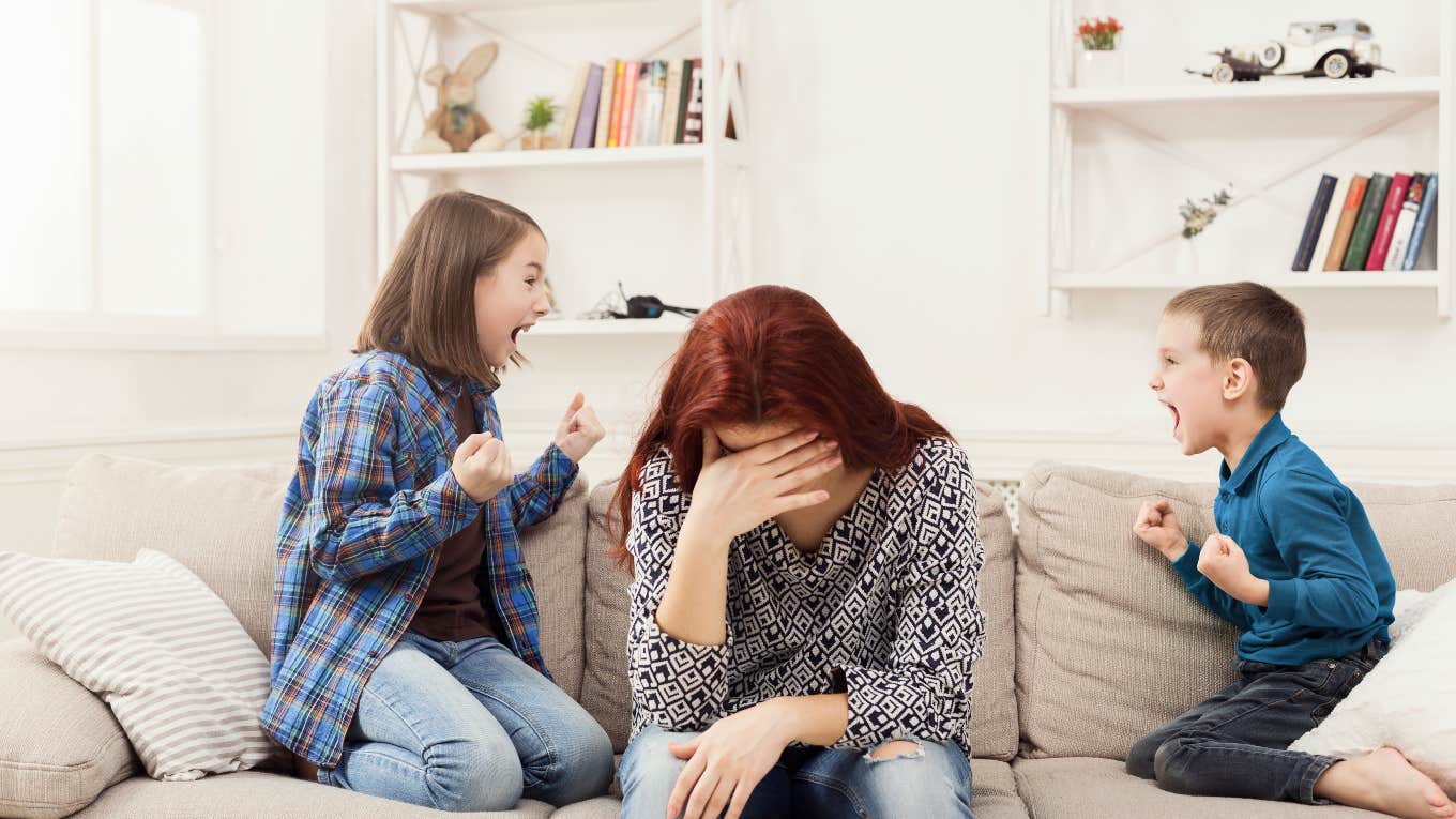 Mom Says It's Harder To Take Care Of Her Kids When Her Husband Is Home two kids fighting while tired, frustrated mom hangs her head between them