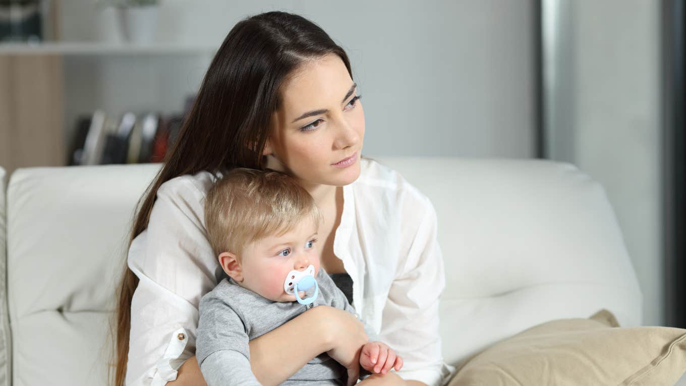 Bride-To-Be Didn’t Invite A Friend To Her Engagement Dinner Because She's A New Parent Mom holding toddler looking sad while sitting on a couch