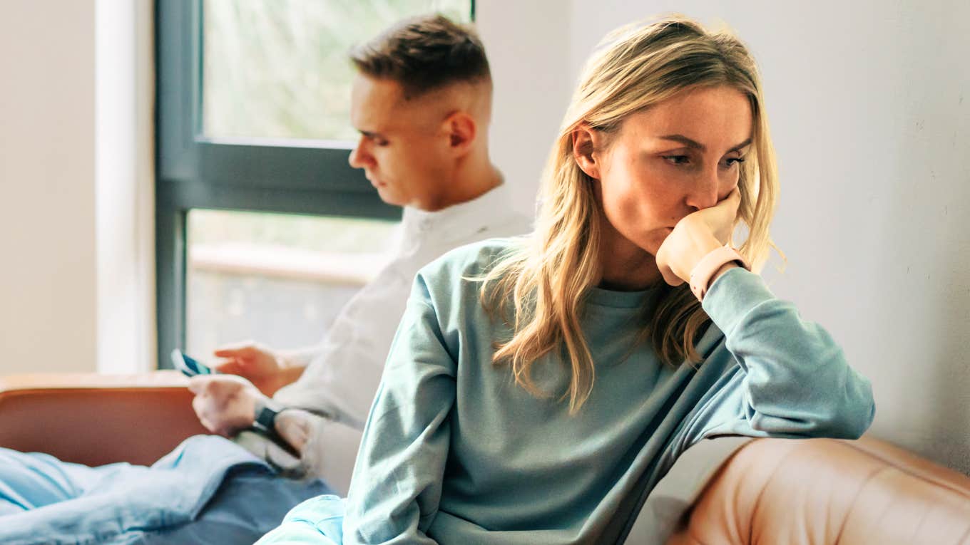 Signs You're Stuck In A Toxic Relationship That Feels 'Fine', According To Experts Stuck in a toxic relationship, reflecting on couch with partner.