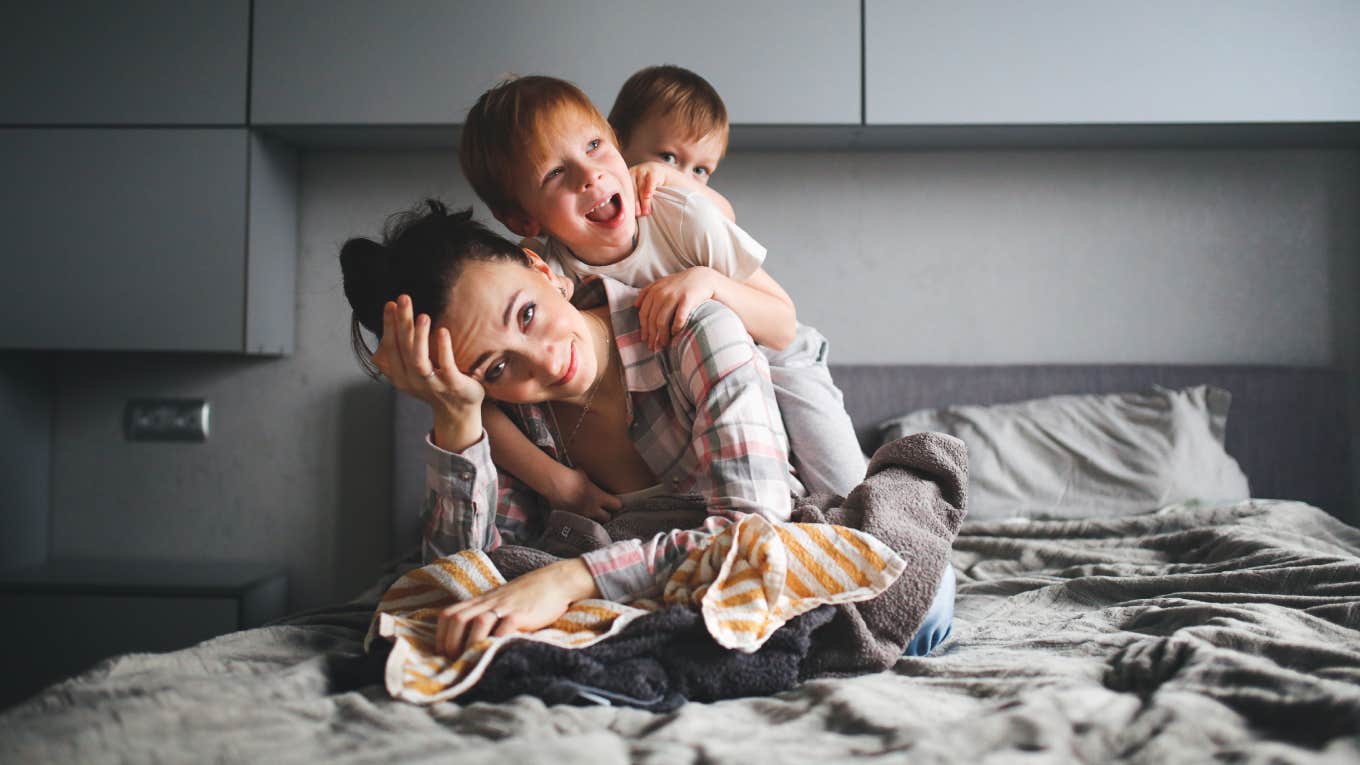 Stay-At-Home Mom Expresses Frustration With Lack Of Validation For The Constant Work She Does tired mother on bed with kids climbing on her back