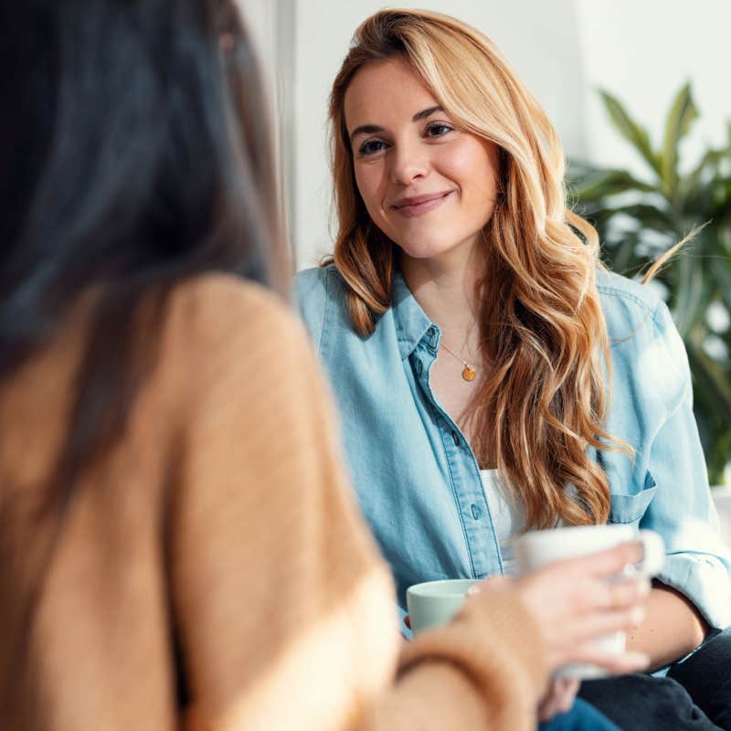 Shot of two smiling young women talking while drinking coffee sitting on couch in the living room at home.