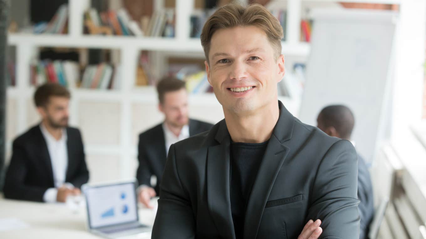 Company Founder Takes Pride In Offering No Work-Life Balance For Employees Smiling confident executive in suit standing with arms crossed looking at camera with business partners team at background
