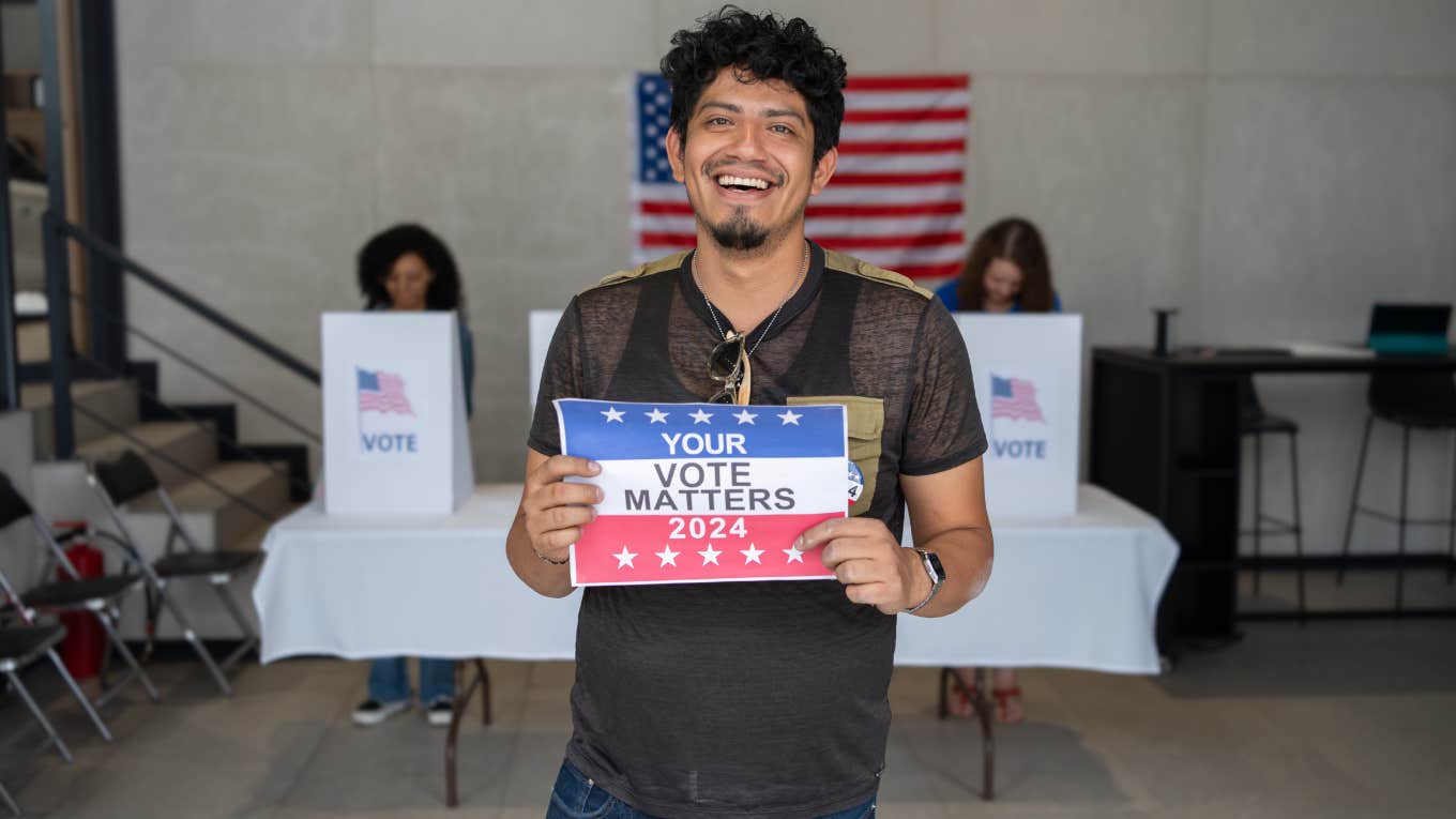 Man Breaks Down While Explaining How It Felt To Vote For The First Time Since Becoming Citizen smiling man holding a Your Vote Matters 2024 sign at a US election polling station