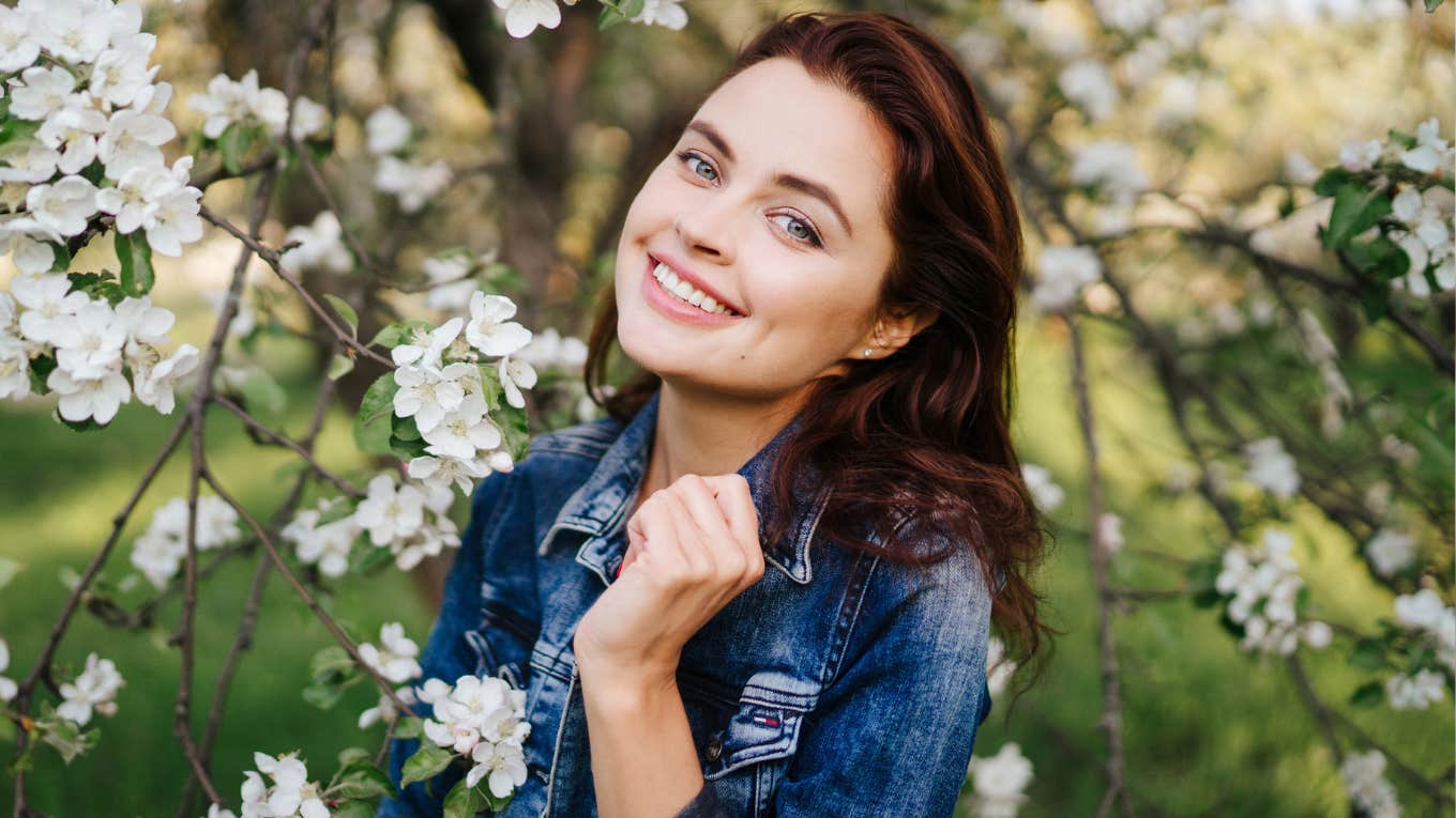 Subtle Traits Of People Who Try Way Too Hard To Impress Everyone Around Them Young smiling woman in jeans jacket standing near blooming spring tree.