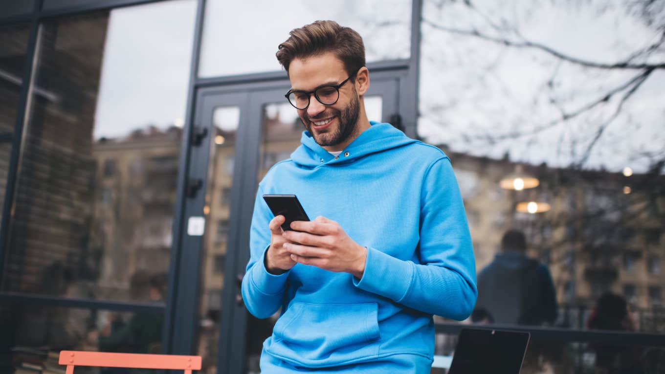 Millennials Are Mourning These Text Acronyms That Are Now Extinct man wearing blue sweatshirt standing outside cafe texting
