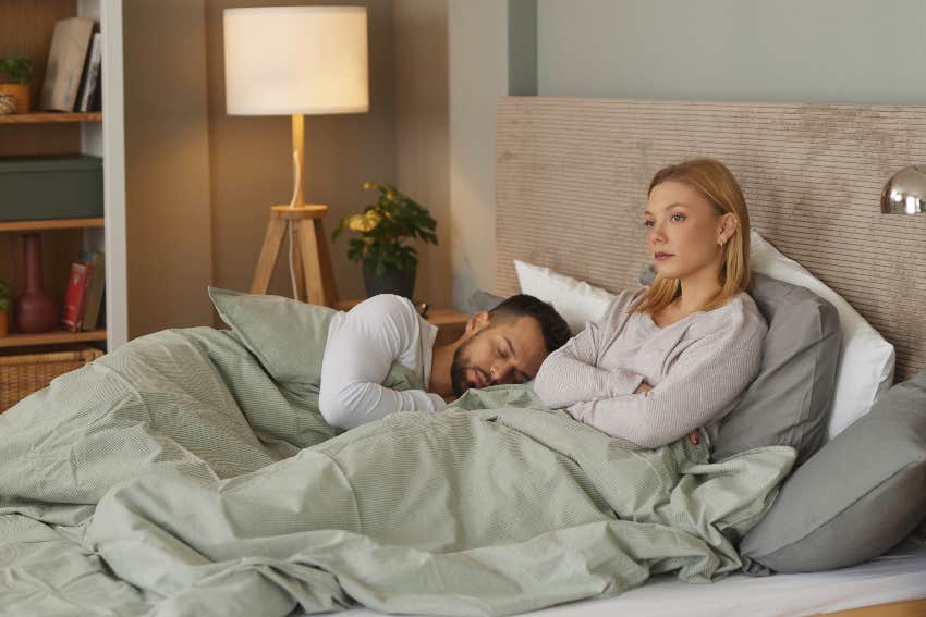 upset woman lying in bed next to sleeping husband