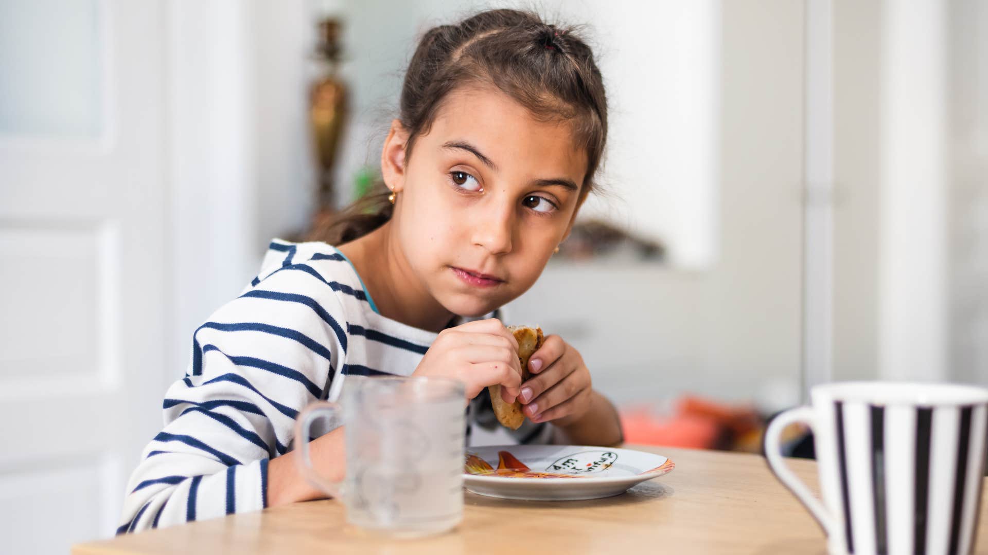Young girl eating a sandwich alone at a table alone instead of having family dinner