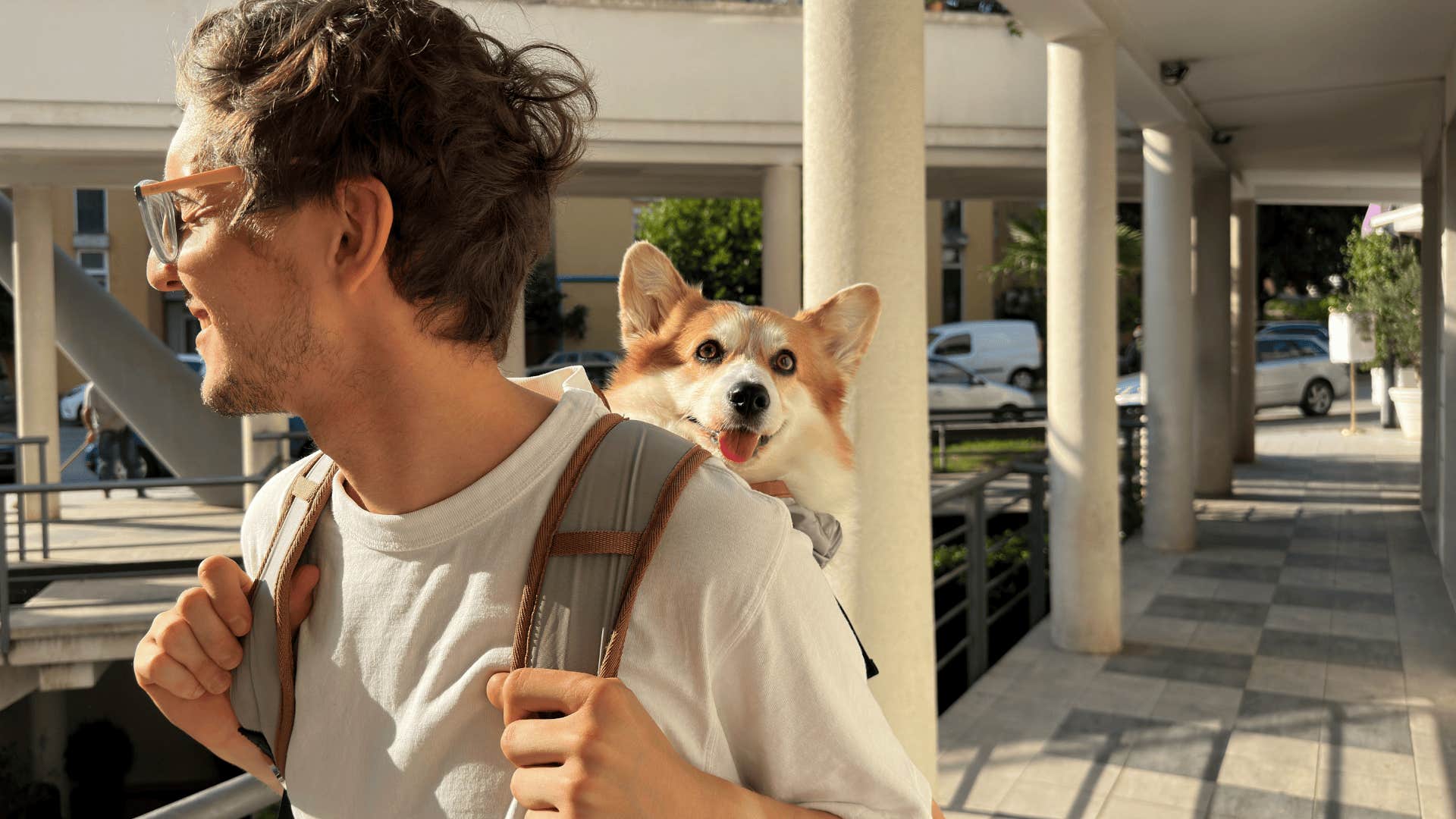 gen z man smiling and traveling with his dog peeking out of a backpack