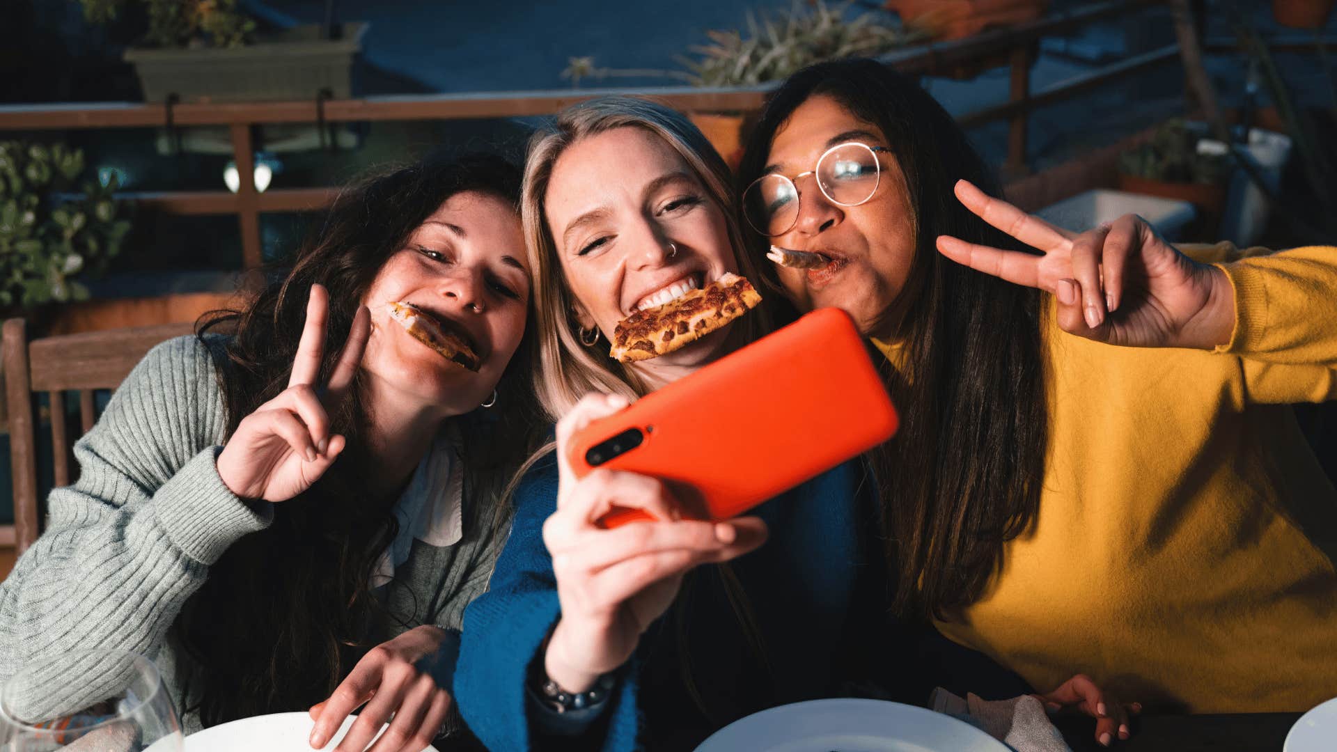 three friends living in the moment as they throw up peace signs and take a picture of themselves with pizza in their mouths