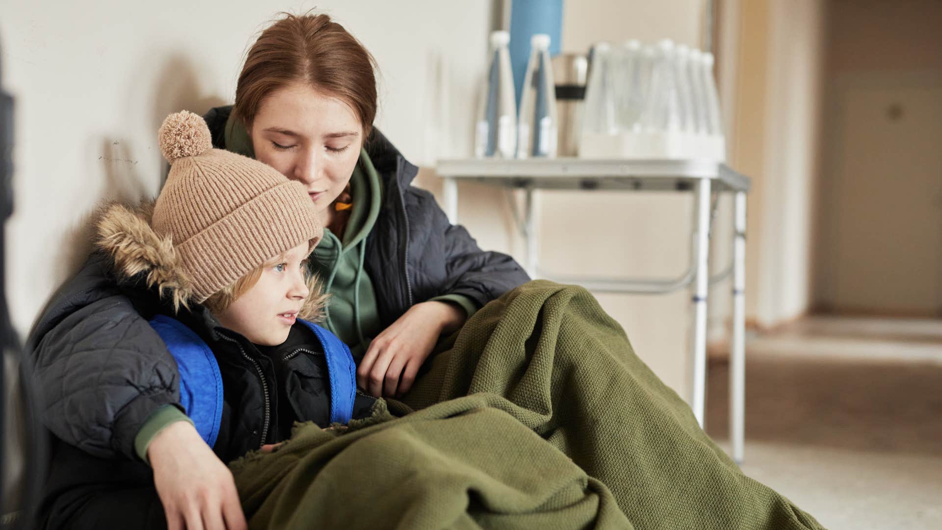 A homeless woman shares her blanket with a child in a shelter.