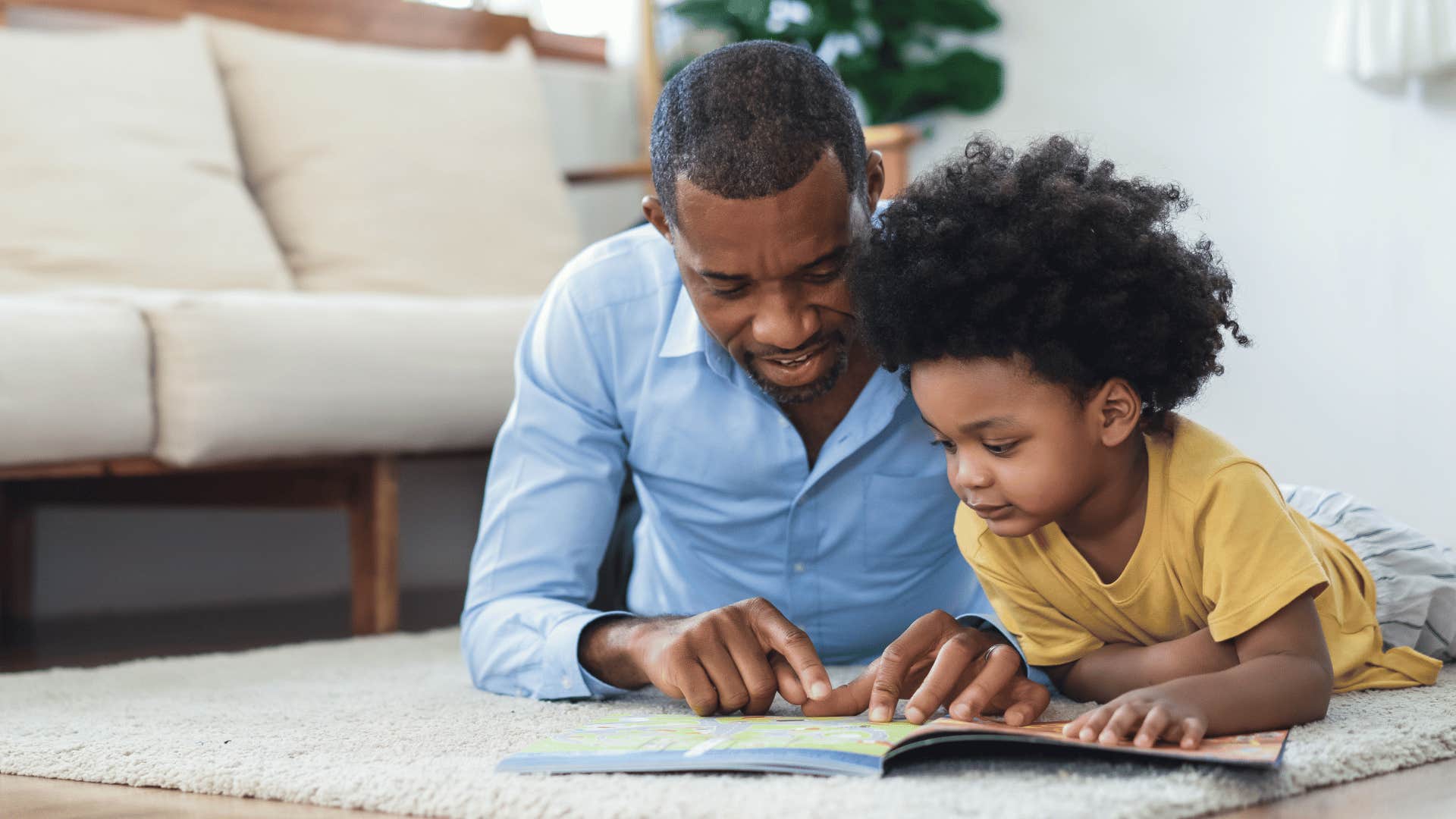 Dad laying on the ground with his son making him read a book.