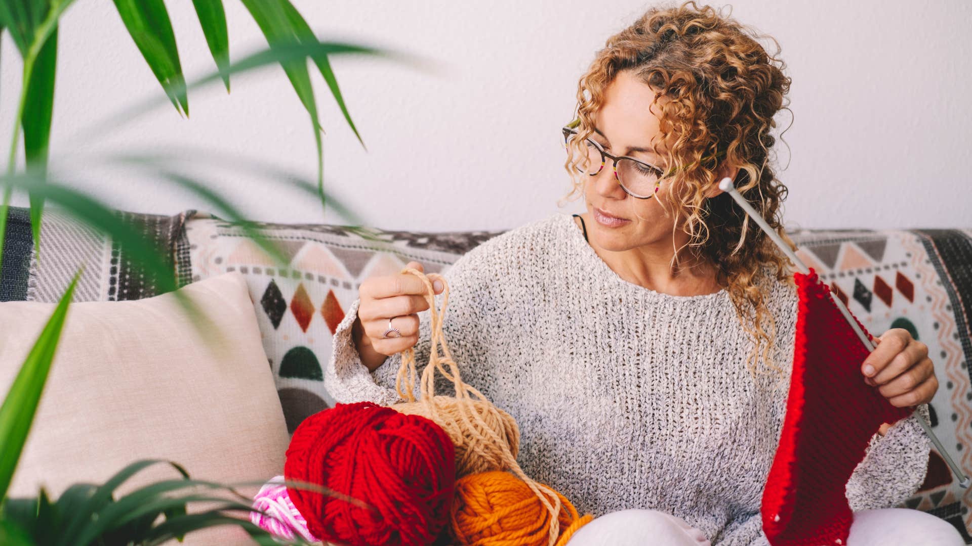 woman in gray shirt enjoying her stay at home hobby of knitting while she knits together red fabric 