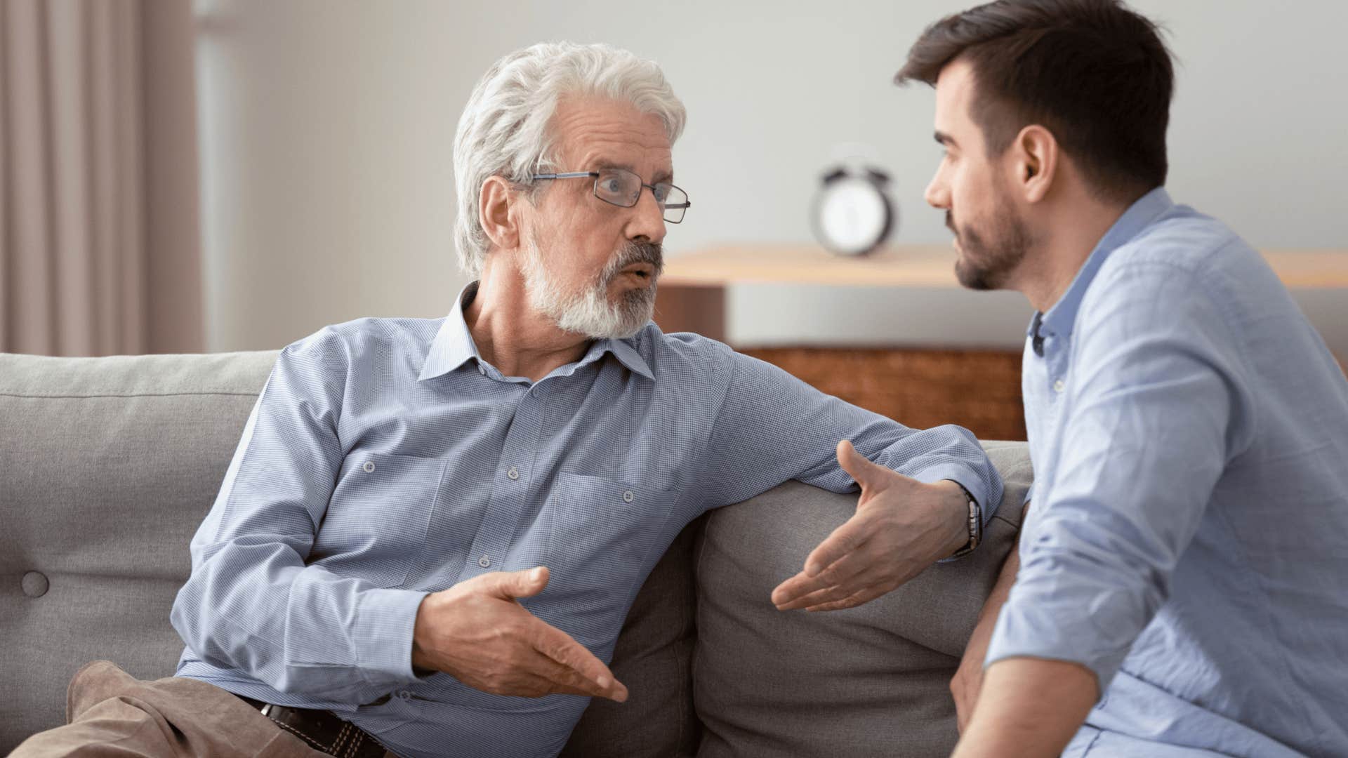 Older serious man saying that’s not my fault to his adult son on the couch