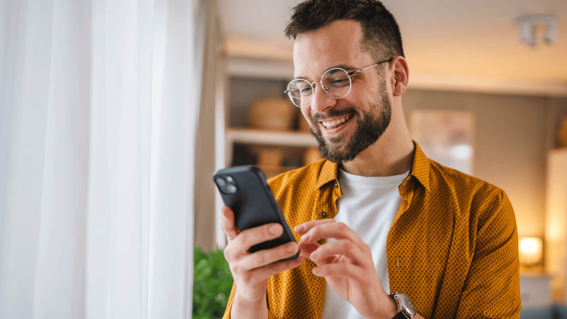 Man chooses his words thoughtfully as he texts on his phone next to a window