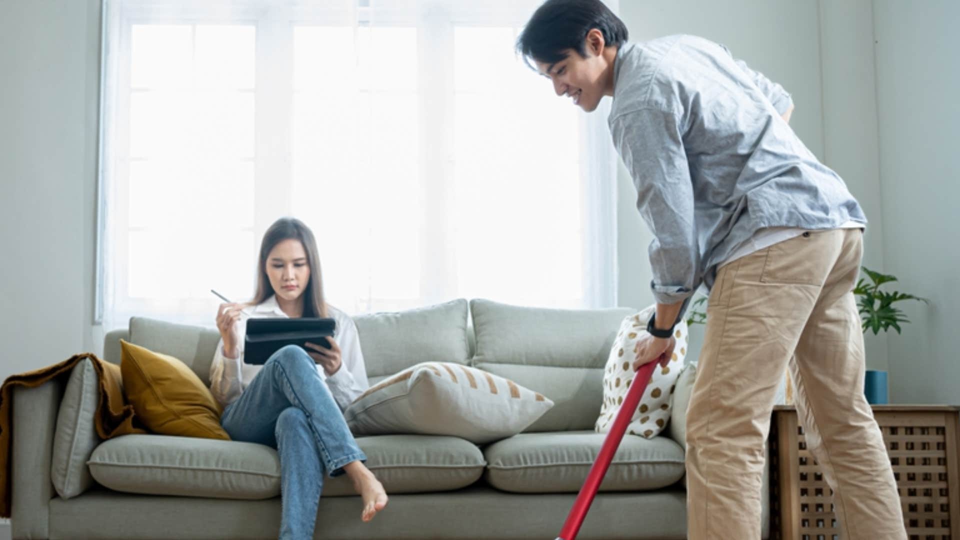 wife sitting on couch while husband does chores in front of her