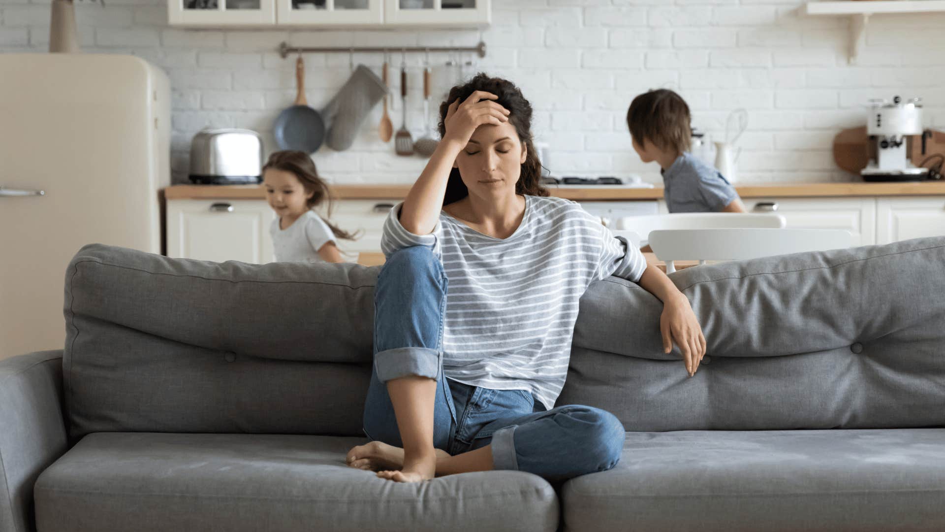 stressed mom sitting on couch with kids running behind her