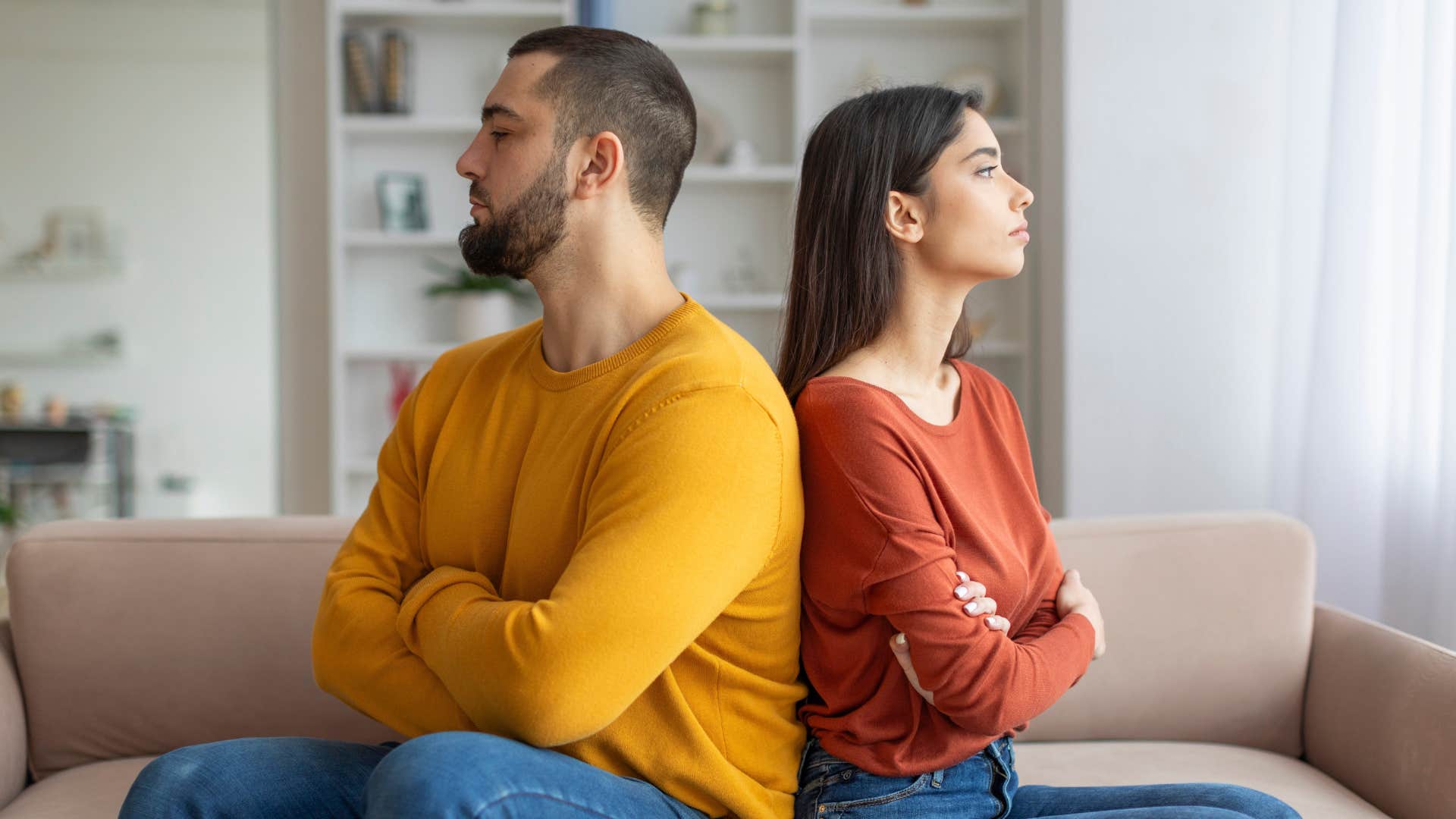 man in yellow-orange shirt and woman in red shirt turned away from each other with arms crossed as they're stonewalling or giving the silent treatment 