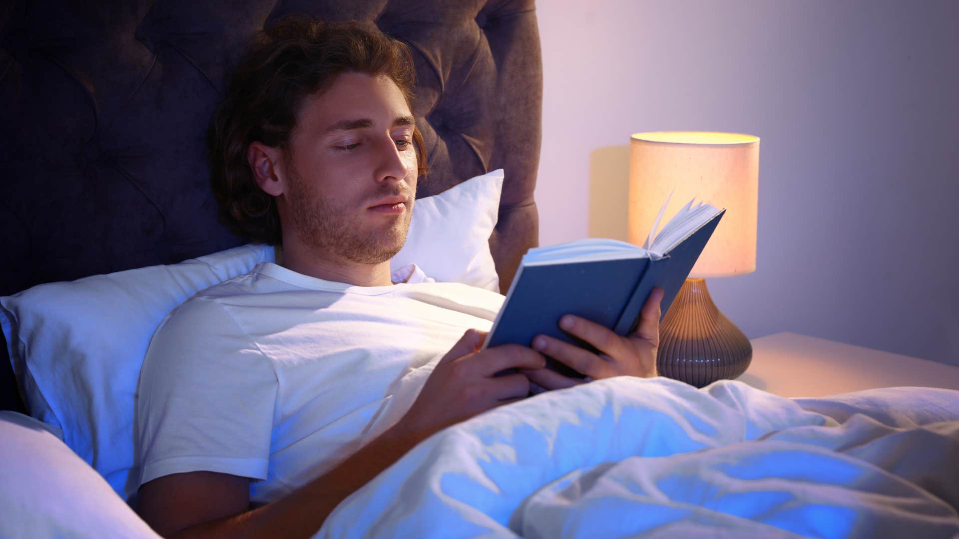 man reading a book in bed with a lamp on
